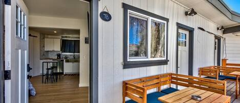 Una terraza de madera con bancos y mesas, un área de cocina visible a través de una puerta abierta, y un letrero con el número 7B en la pared.