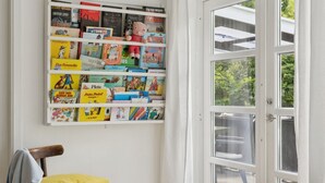 Een kamer met een wit wandplankje vol kinderboeken, een houten stoel met een geel kussen, en een witte tafel.