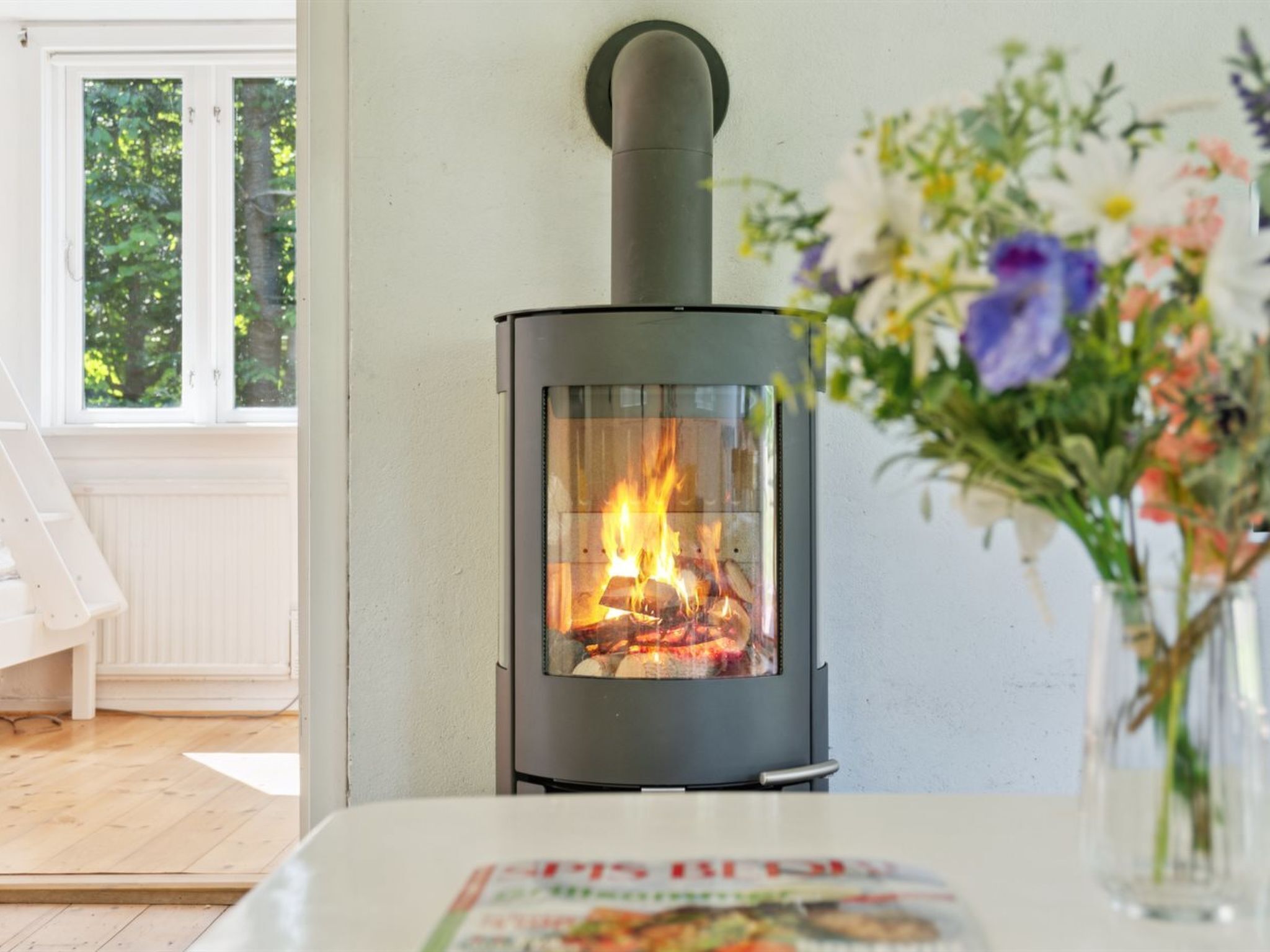 A wood-burning stove in a room with a white table, a vase of flowers, and a window with a view of trees.