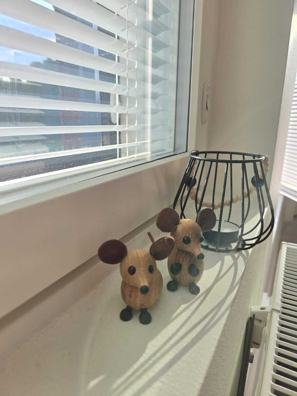 A window with closed blinds, a small metal cage, and two wooden dog figurines on a white surface.