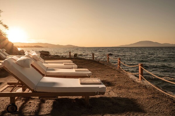 A beachside lounge area with sunbeds, a rope barrier, and a view of the sea and distant mountains.
