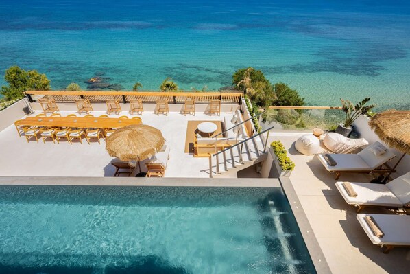 An infinity pool with clear blue water, a sun terrace with lounge chairs and a dining table, and a view of the ocean.