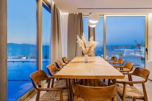 A modern dining area with a wooden table and chairs, large windows with a view of the sea, and a vase with pampas grass.