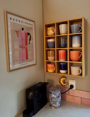 Una cocina con cafetera, un cuadro enmarcado y una repisa con diferentes tazas.