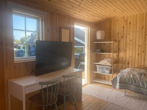 A room with wooden walls, a white desk with a TV, two chairs, a bed, and a window.