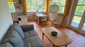 A living room with a sofa, a dining table with chairs, a kitchen area, and large windows with curtains.