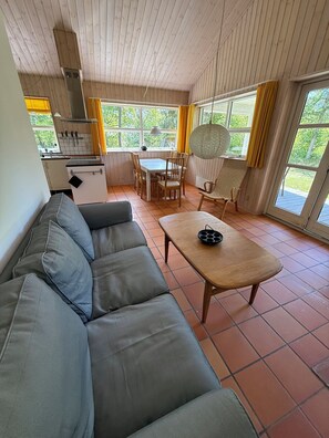A living room with a sofa, a dining table with chairs, a kitchen area, and large windows with curtains.