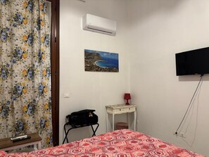 A bedroom with a bed, a painting of a coastal scene, an air conditioner, a TV mounted on the wall, and a desk with a lamp.