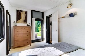 Et soveværelse med seng, kommode, et vindue med gardiner og en vægmonteret lampe.
