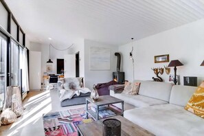 A modern living room with a sofa, coffee table, and a fireplace. There is a kitchen in the background, and the room has large windows.