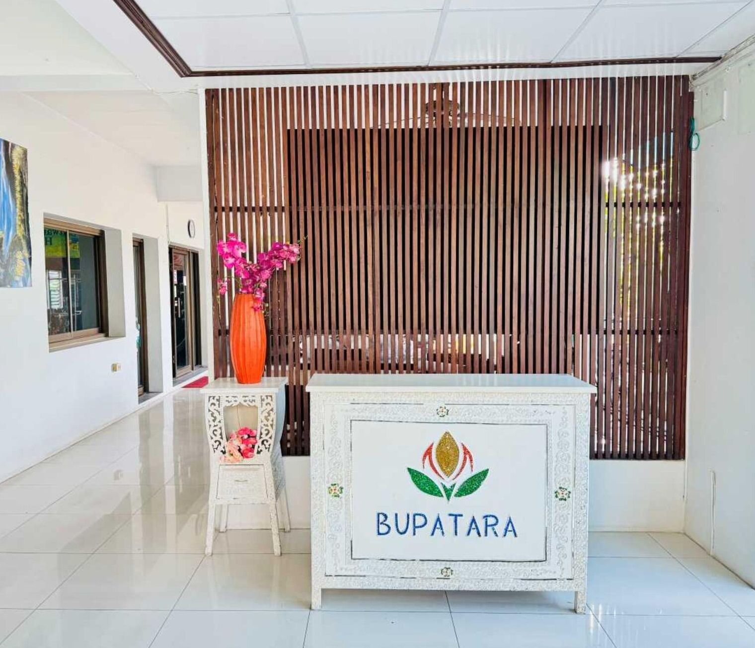 A reception area with a white reception desk labeled "BUPATARA," a small table with a vase of pink flowers, and a wooden slatted wall in the background.