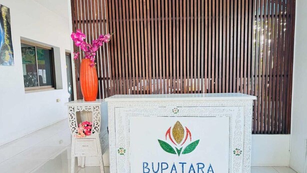 Una recepción con el nombre 'BUPATARA' y un logo, una pequeña silla blanca, un jarrón con flores rosadas y un fondo de listones de madera.