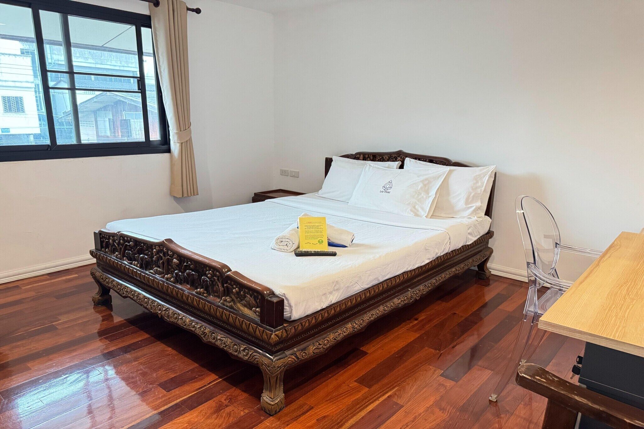 A neatly made bed with a wooden frame and white linens, a bedside table with a lamp, and a window with curtains.