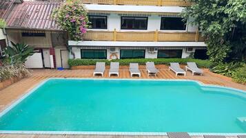 A swimming pool with lounge chairs, a building with balconies, and a tiled walkway.