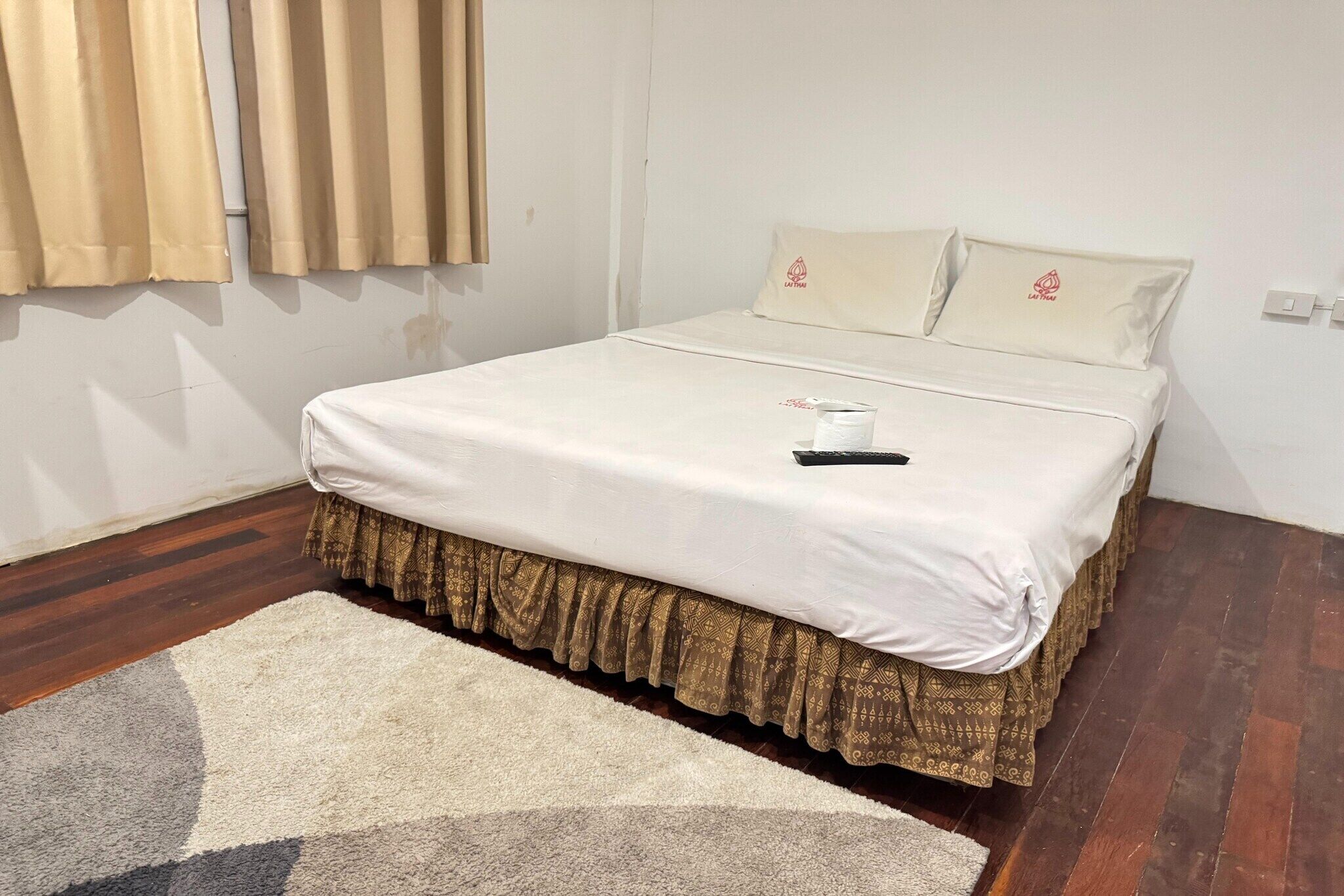 A neatly made bed with a patterned skirt, a white pillow with a logo, and a remote control on the bed.