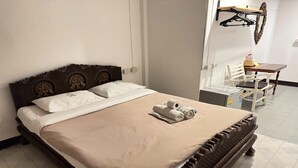 A neatly made bed with a beige bedspread and two pillows, a wooden headboard with decorative carvings, and a small table with a chair in the background. - Budget Double Room | Simple & Comfy (Chiang Mai)