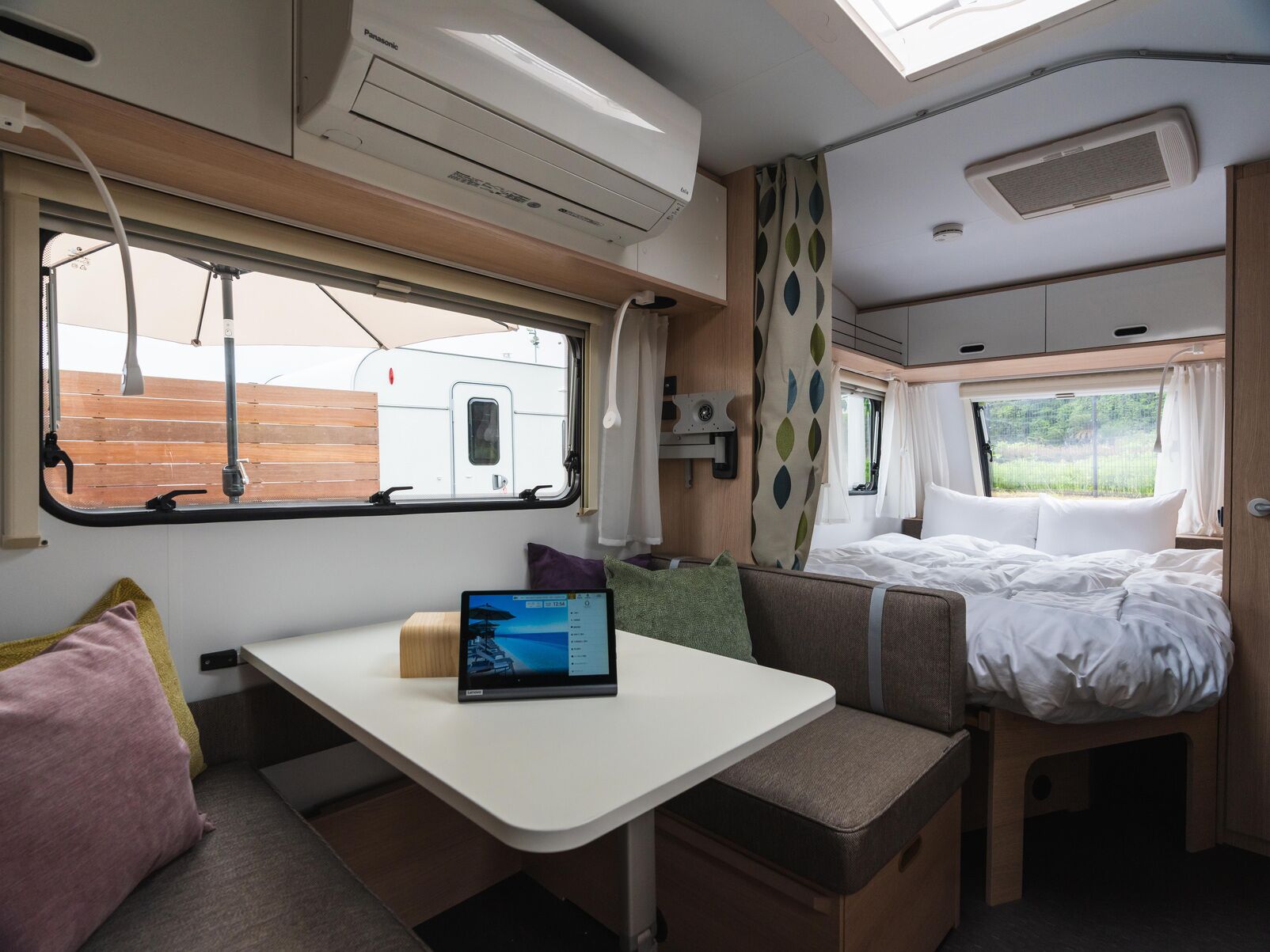 A compact RV interior with a bed, a small table with a laptop, and a window overlooking a wooden deck.
