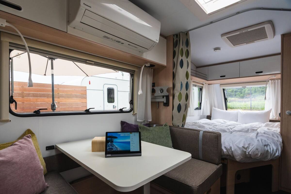A compact RV interior with a bed, a small table with a laptop, and a window overlooking a wooden deck.