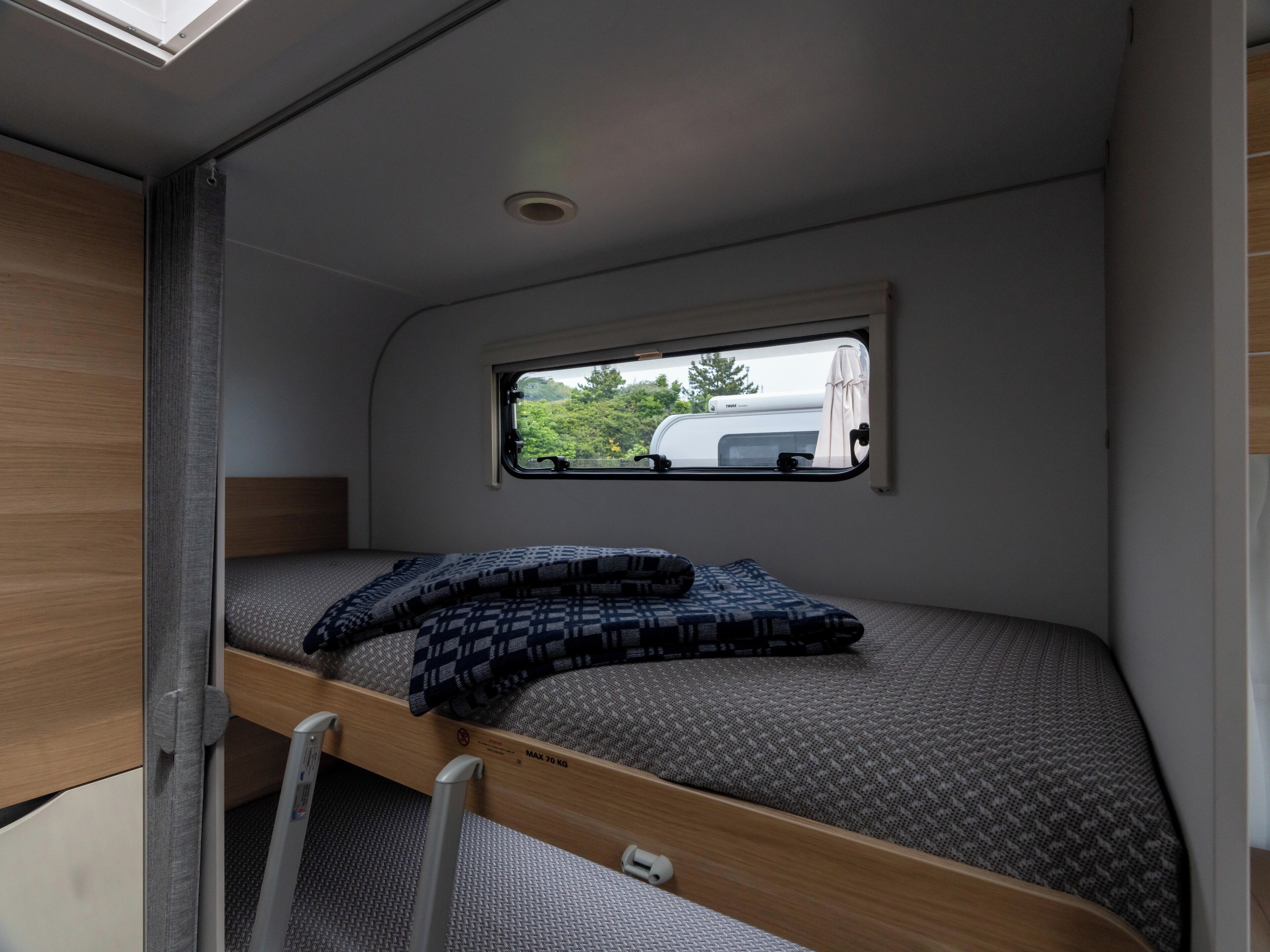 A compact RV interior with a bunk bed, a window with a view of a white RV, and a skylight above.