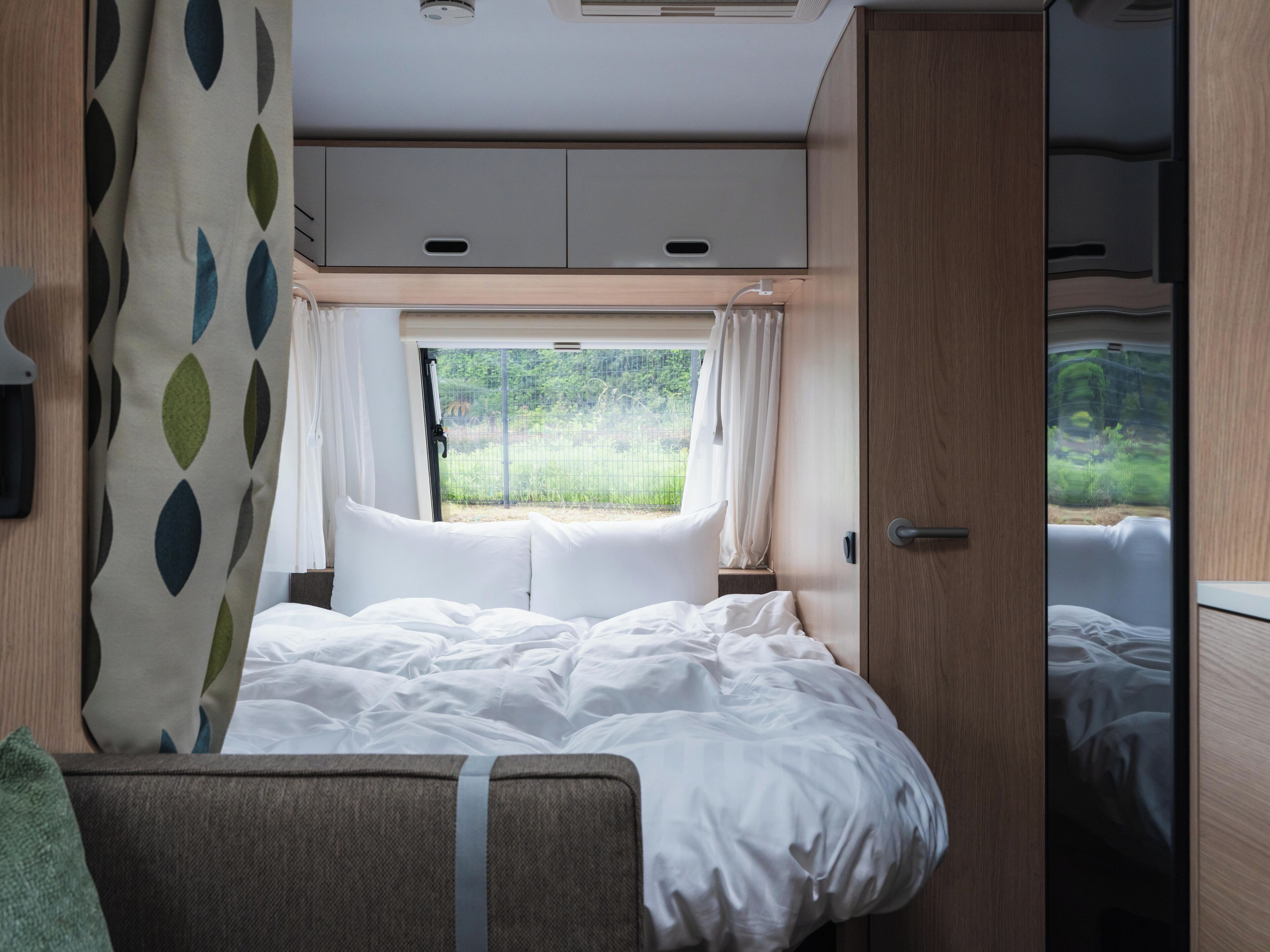 A cozy RV bedroom with a bed, a window with curtains, and a view of greenery outside.