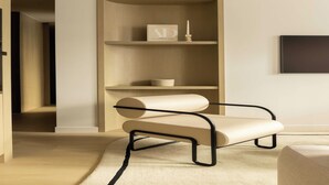 Una habitación moderna y minimalista con suelo de madera, un banco color beige con estructura de metal negro, un estante de madera con un libro y una vela, y un televisor de pantalla plana montado en la pared.