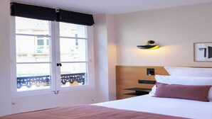 Habitación de hotel con cama, mesita de noche, lámpara de pared y ventana con vista a un balcón.