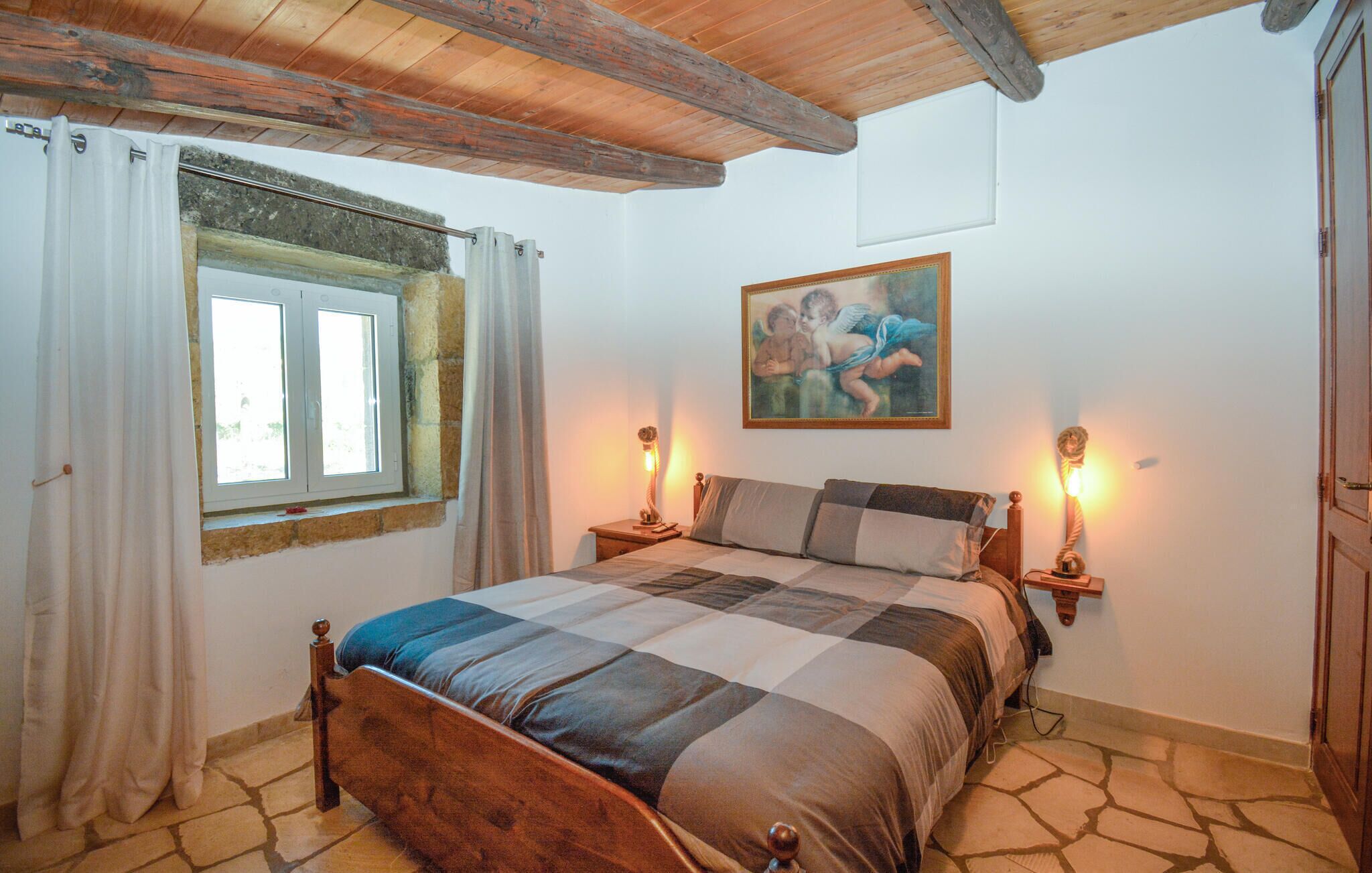 A bedroom with a wooden bed, stone flooring, a window with curtains, and two bedside lamps.