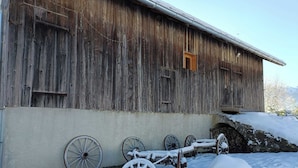 Un granero de madera rústico con una sola puerta y ventanas, rodeado de nieve y ruedas de carro antiguas y oxidadas.