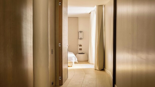 A hotel corridor with wooden doors, a bed with white bedding, a bedside table, and a wall-mounted light fixture.