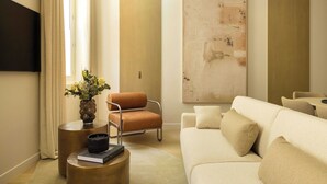 A modern living room with a beige sofa, a brown armchair, a round side table with a book and a candle, and a large abstract painting on the wall.