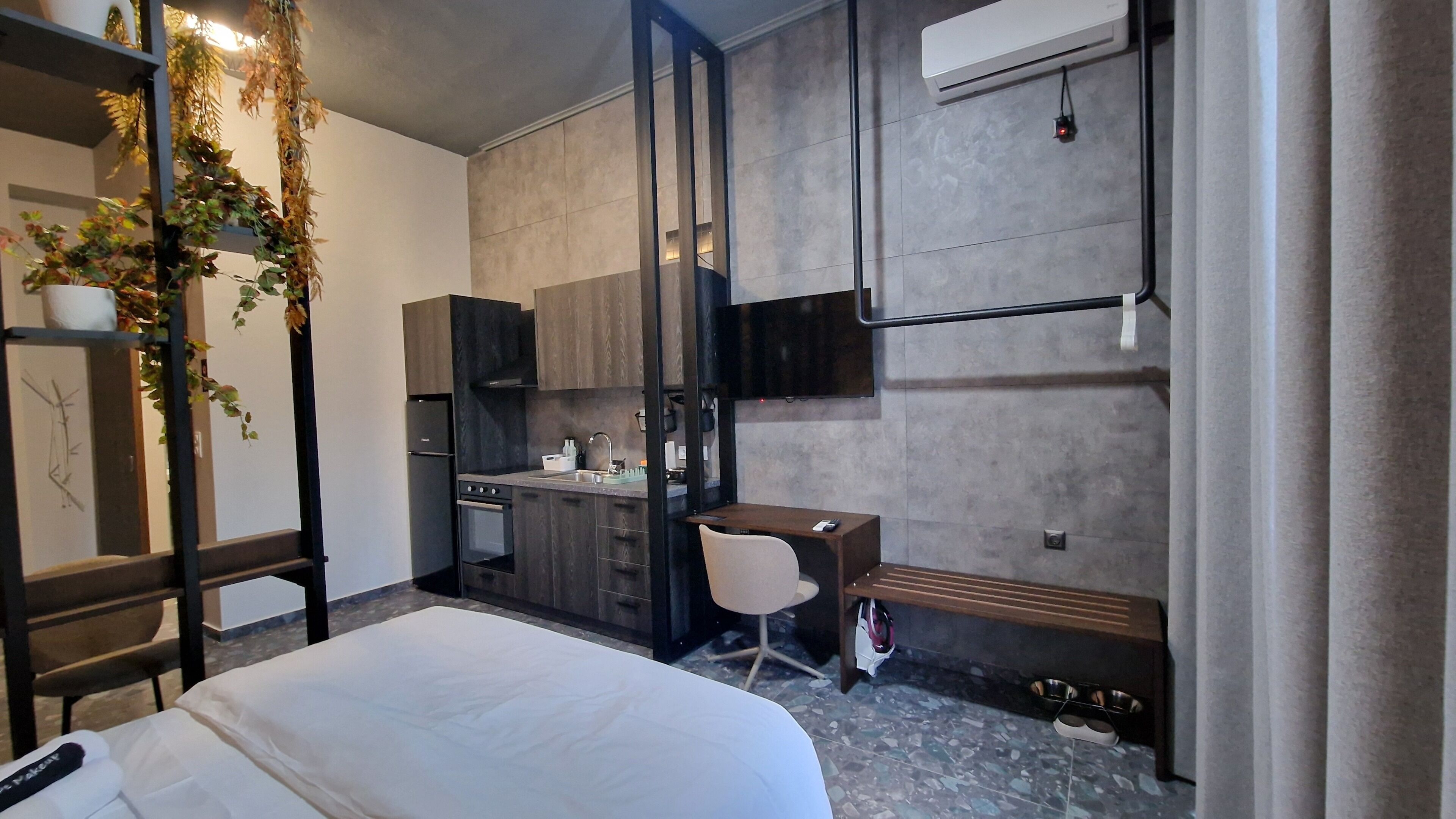 A modern hotel room with a bed, a desk, a chair, a kitchenette, and a TV mounted on the wall.