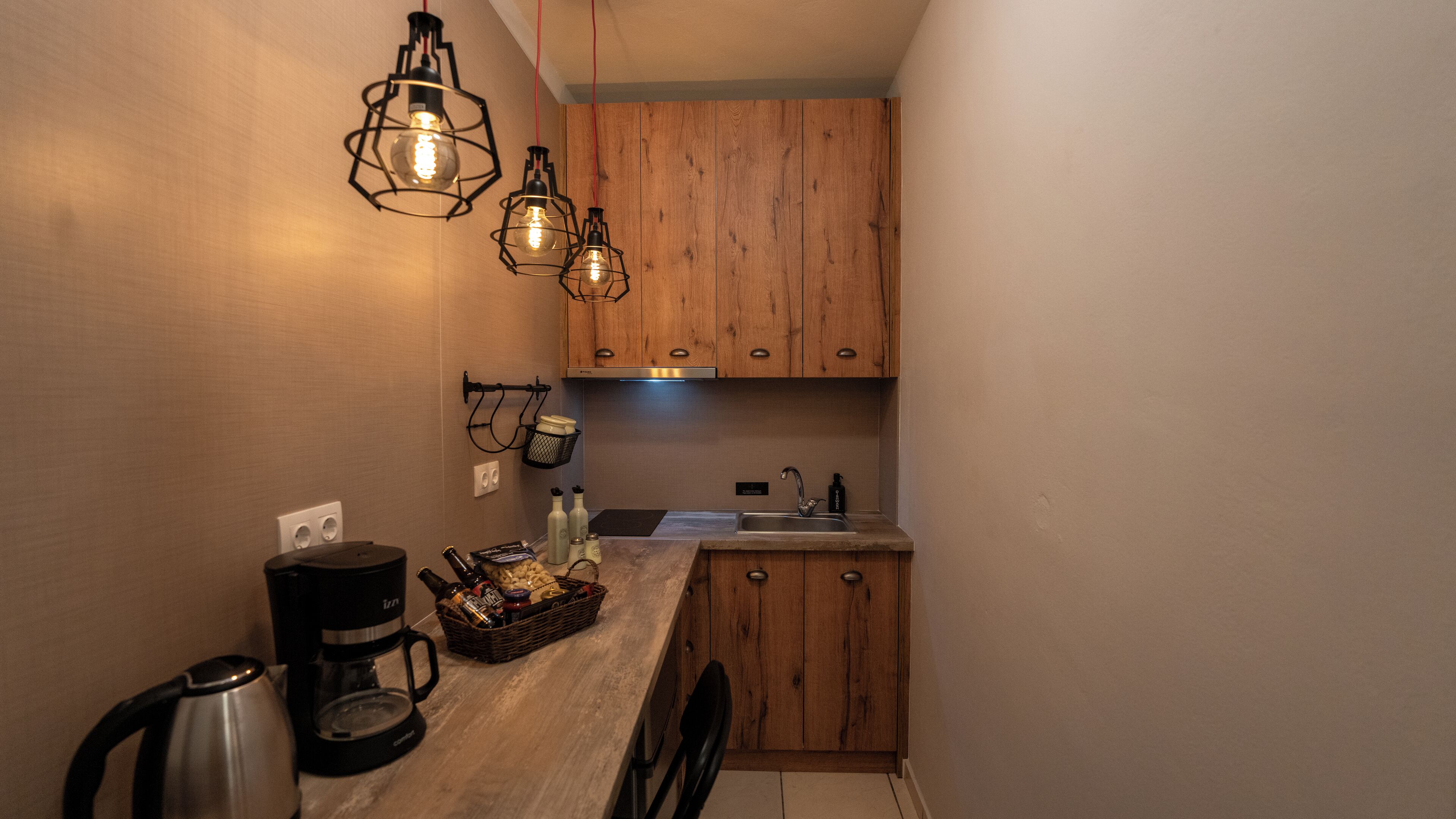 A compact kitchen with wooden cabinets, a coffee maker, a kettle, and two pendant lights.