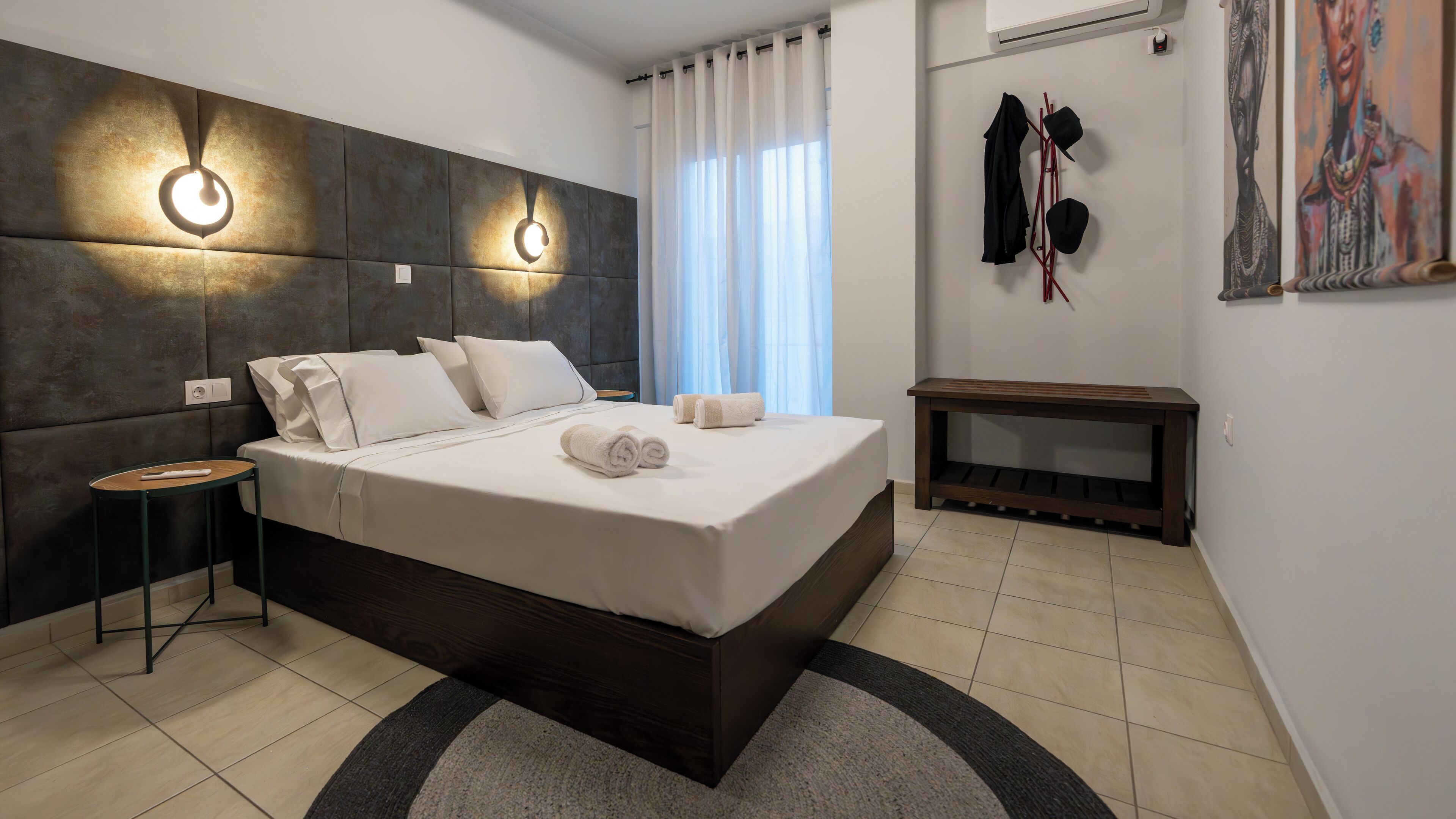 A modern bedroom with a large bed, two wall-mounted lights, a bedside table, a small bench, and a painting on the wall.
