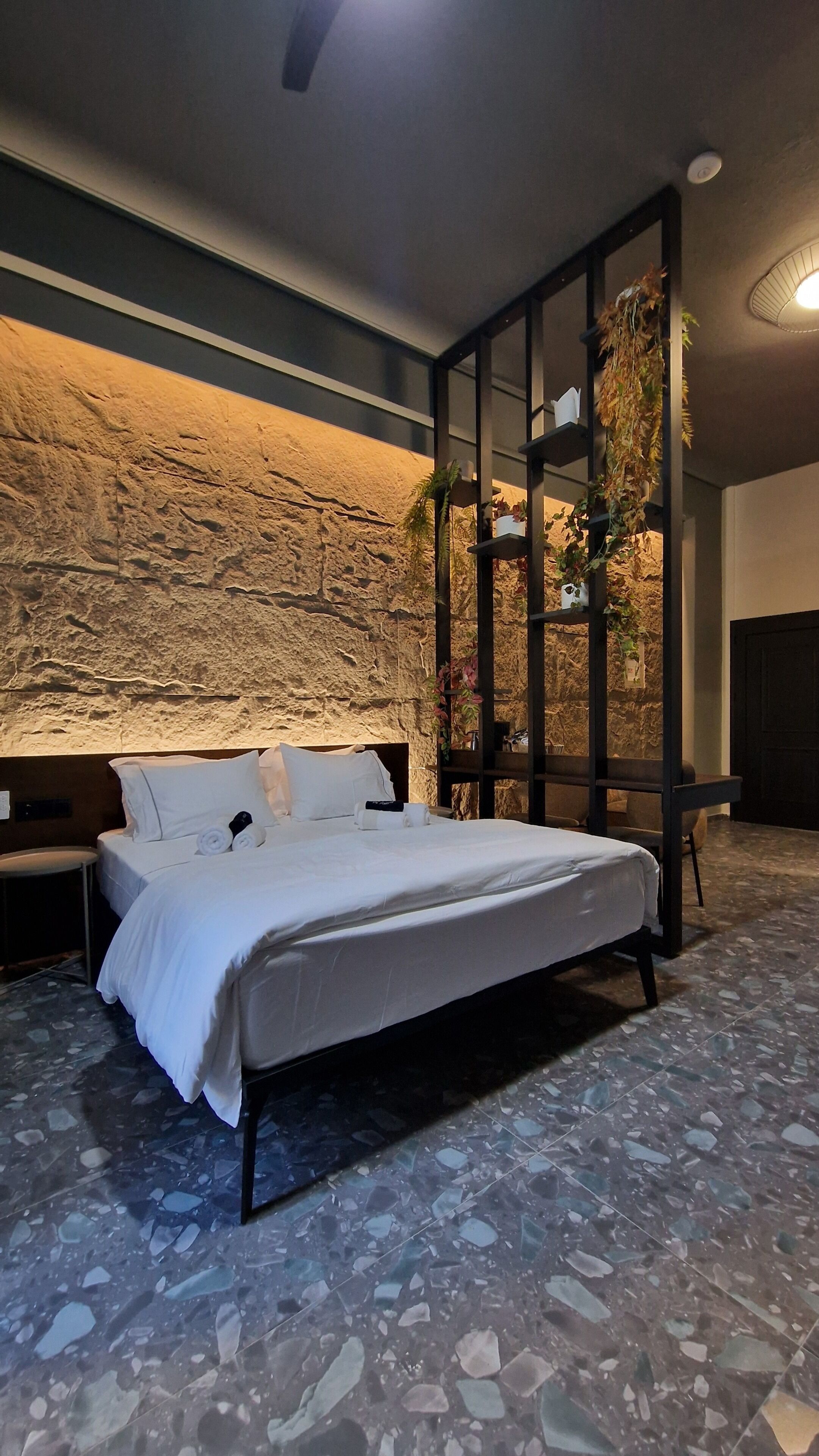 A bedroom with a stone wall, a bed with white linens, a nightstand, and a large window with a view of greenery.