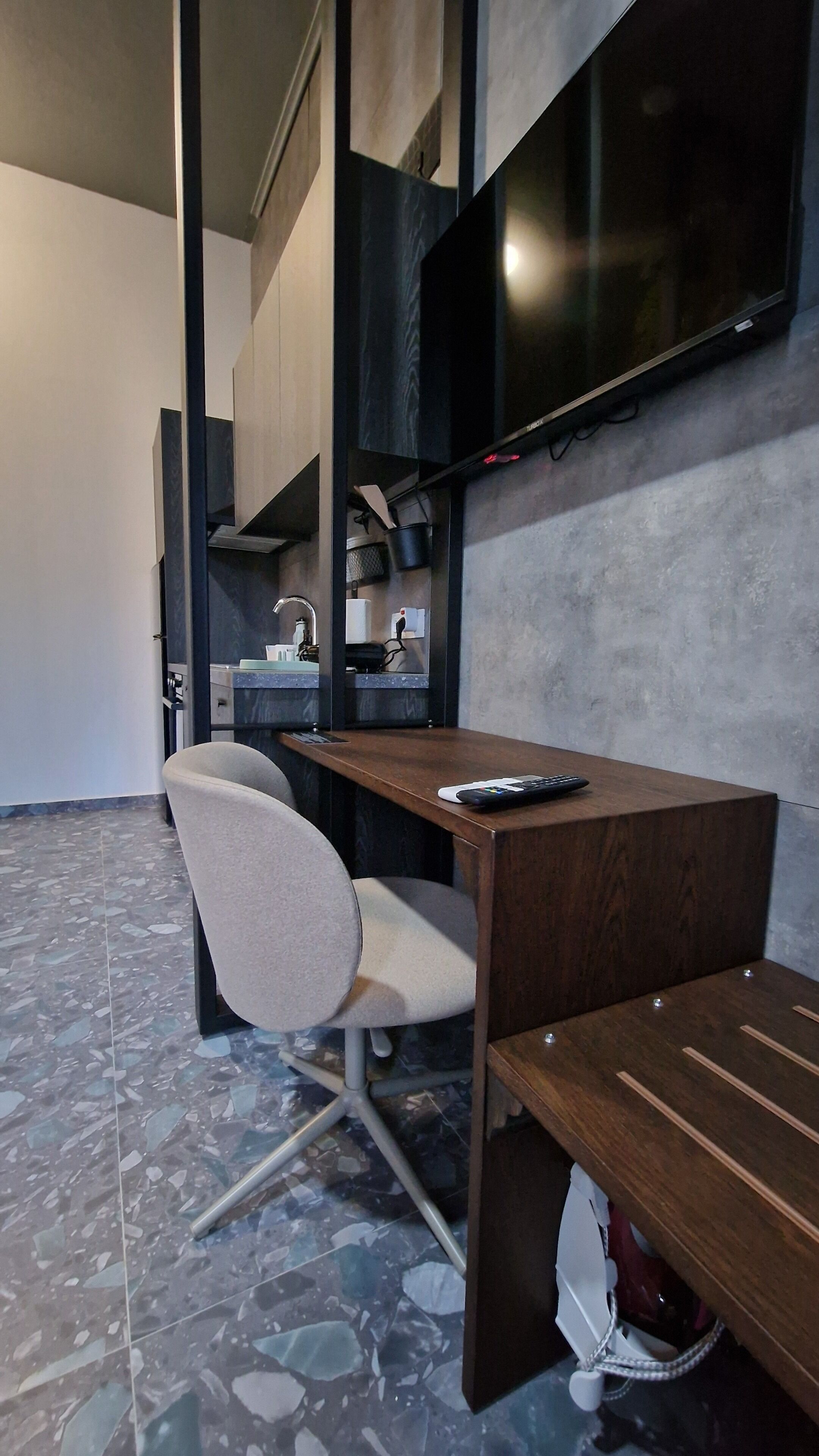 A modern room with a wooden desk, a chair, a TV mounted on the wall, and a kitchen area with cabinets and a sink.