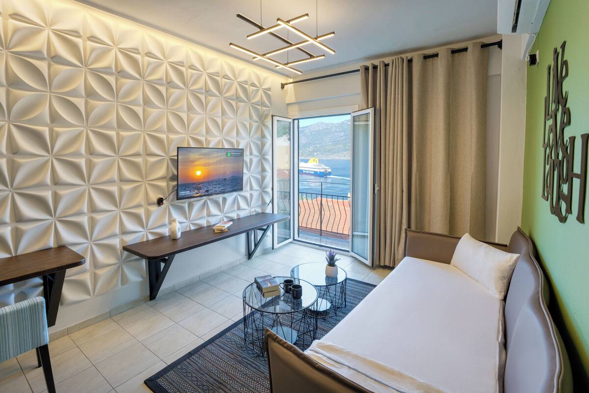 A modern hotel room with a geometric patterned wall, a flat-screen TV, a bed, a desk, and a view of the sea through a large window.
