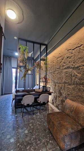 A modern interior with a stone wall, a round ceiling light, a dining area with chairs, a sofa, and potted plants. - WELL BEING LUXURY APARTMENTS (Ikaria)