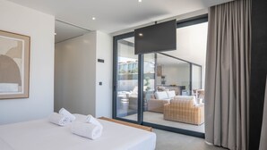 Quarto de hotel moderno com uma cama grande, televisão de tela plana e uma porta de vidro deslizante que dá para uma varanda com mobiliário de vime.