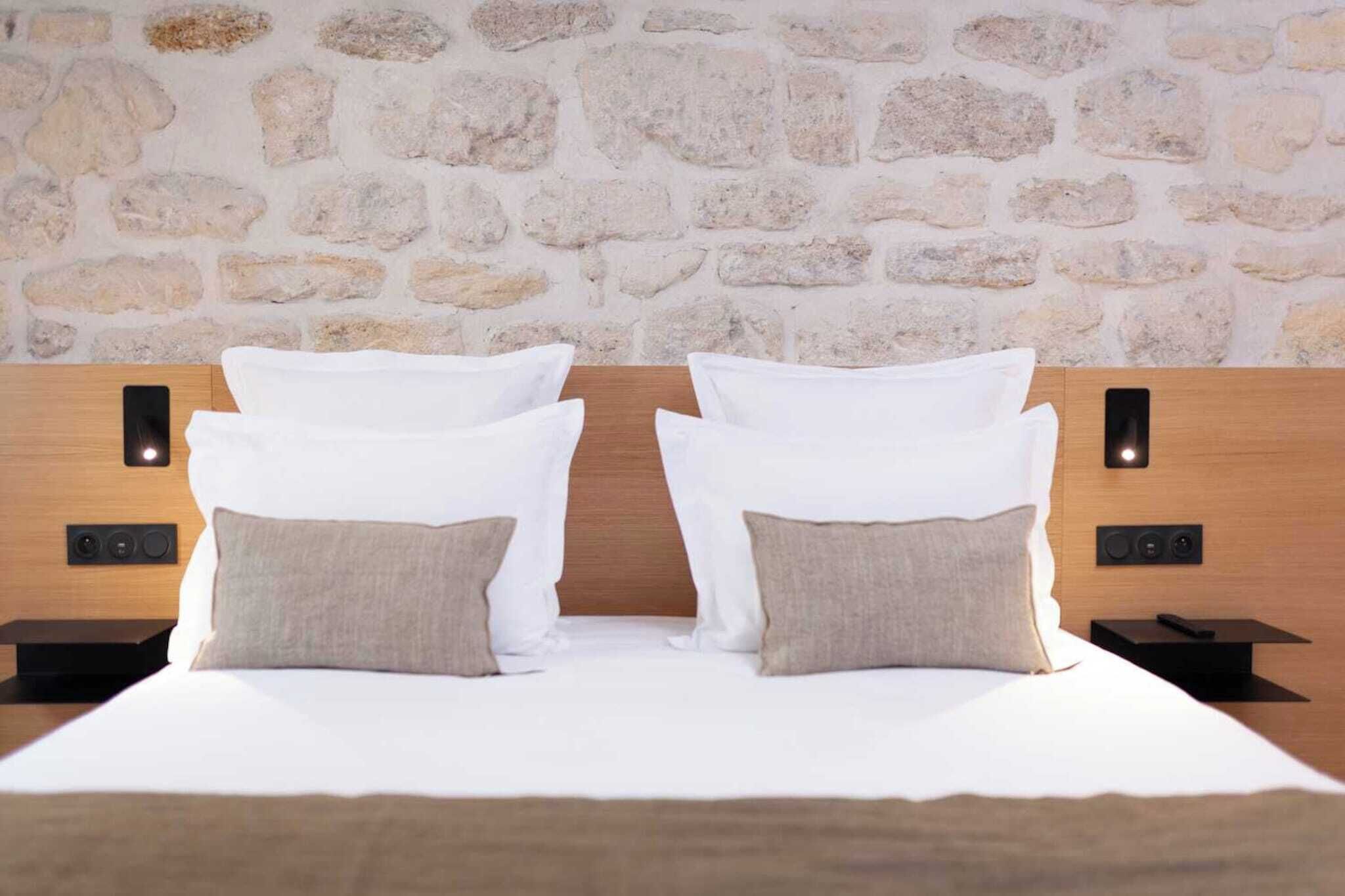 Una cama doble con almohadas blancas y beige, un cabecero de madera y dos mesitas de noche con lámparas de pared.