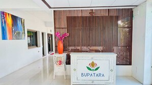 A reception area with a white reception desk labeled "BUPATARA," a wooden slat wall, a colorful abstract painting, and a small decorative table with pink flowers. - Superior Double Stay | Sleek & Cozy (Chiang Mai)