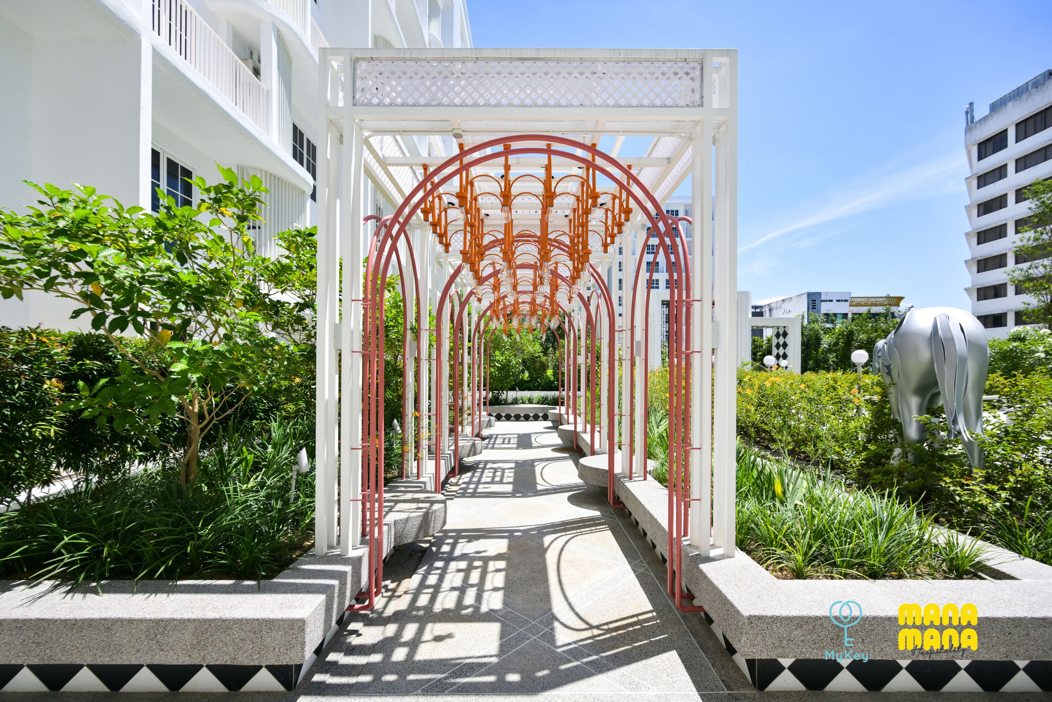 A pathway leading to a white building with a red metal archway, surrounded by greenery and a large statue.