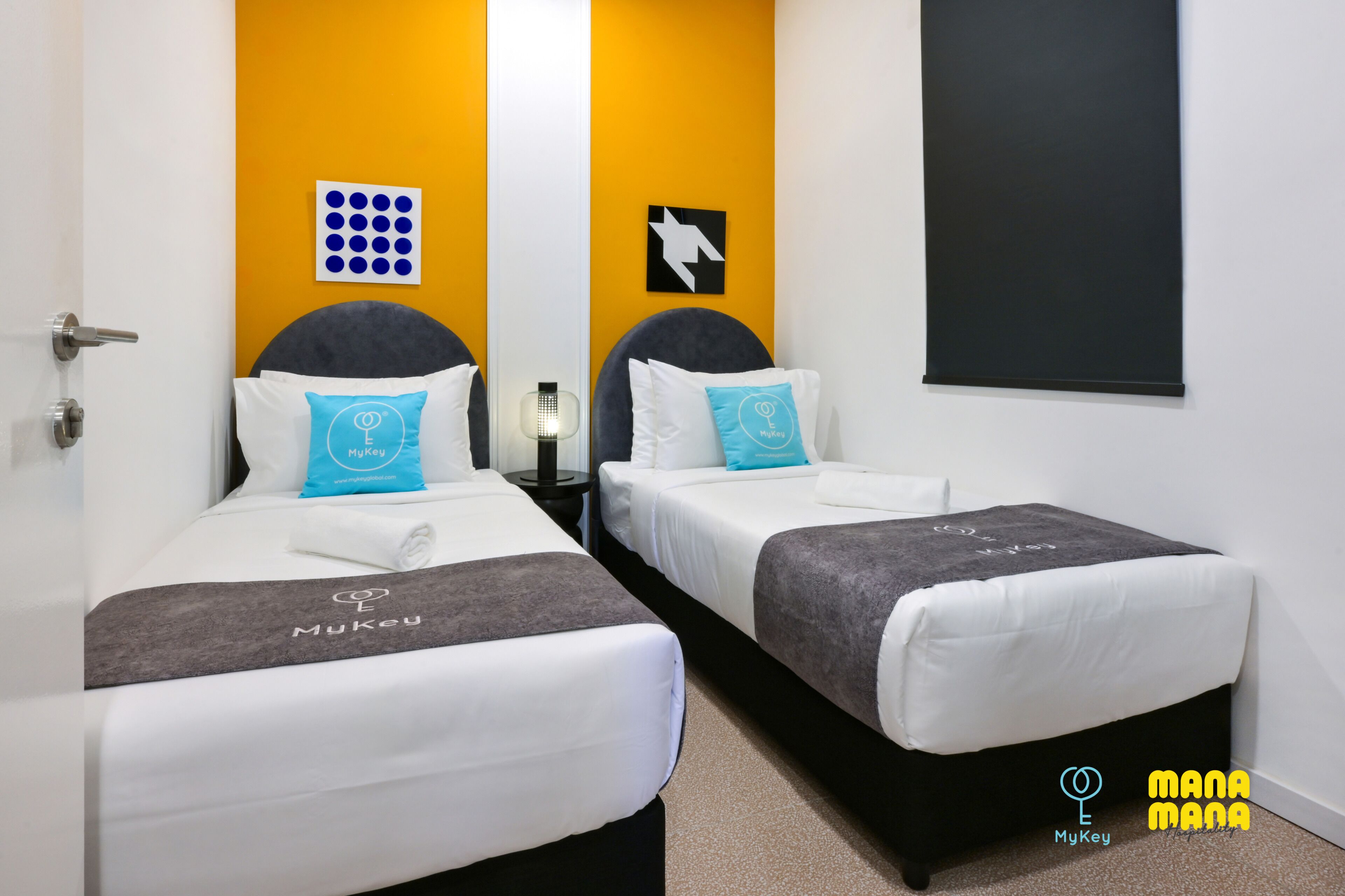 Ein Hotelzimmer mit zwei Betten, jedes mit grauem Kopfteil und weißer Bettwäsche. Es gibt zwei Kissen mit dem Logo des Hotels, eine wandmontierte Lampe und eine Tafel an der Wand.