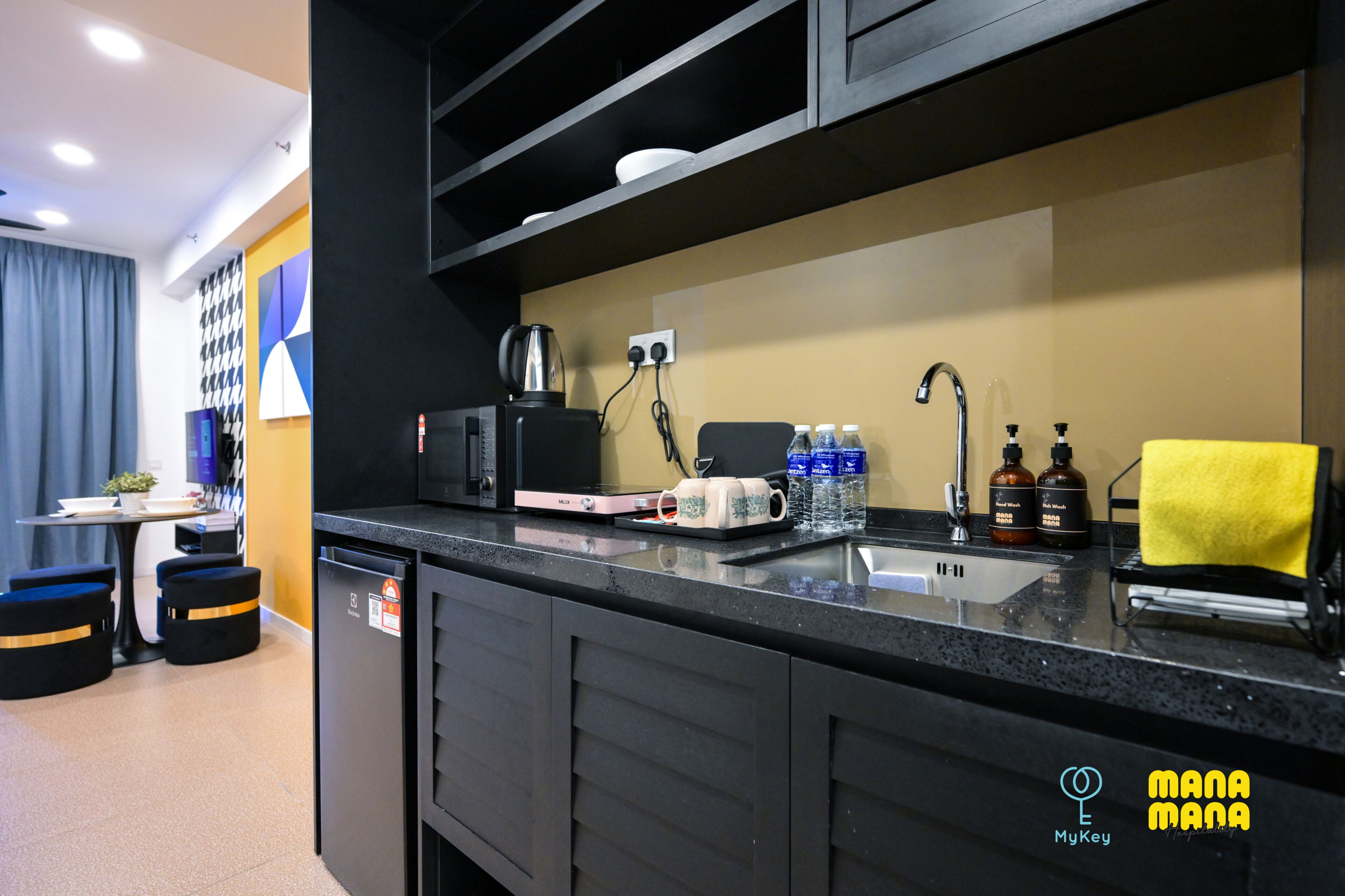 A modern kitchen with a black countertop, a coffee maker, a kettle, and a sink. There are two stools and a small table with a plant.