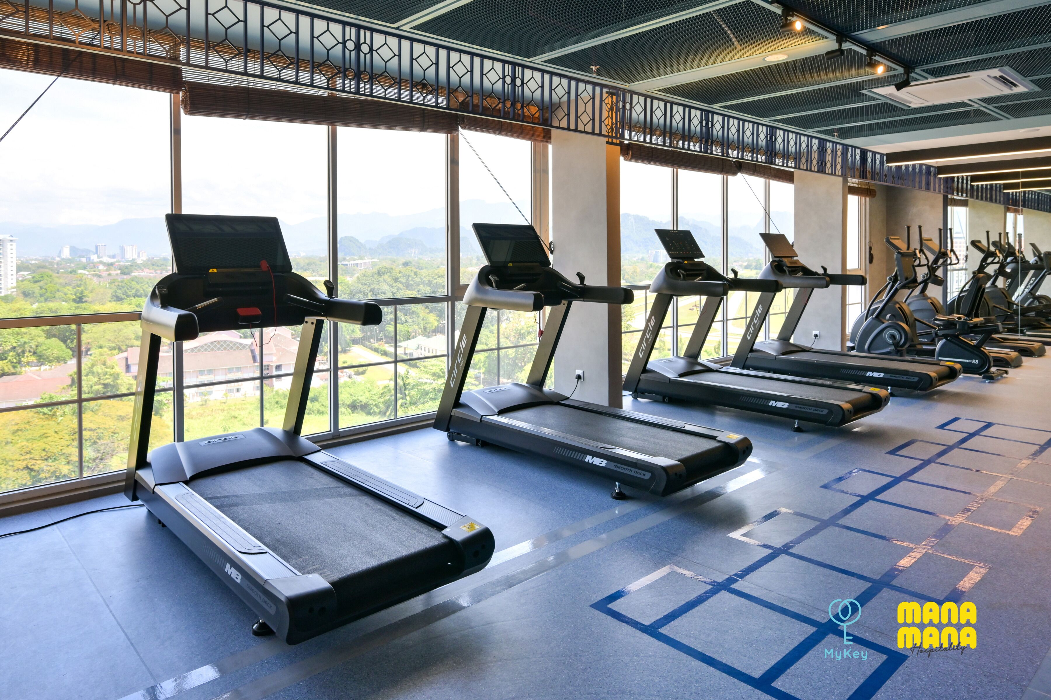 A gym with treadmills aligned in rows, large windows offering a view of the cityscape, and a blue patterned floor.