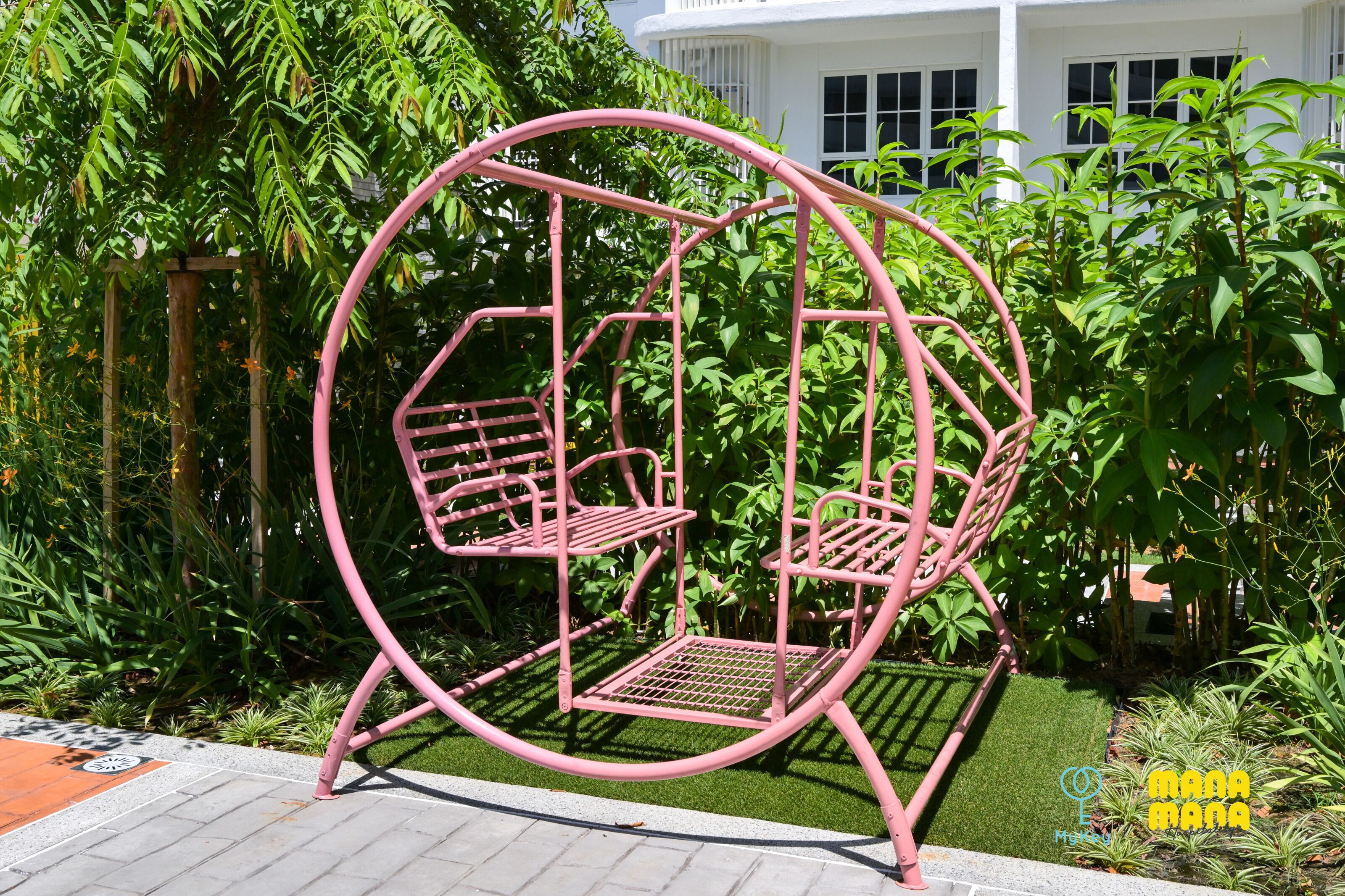 A pink swing set in a garden with green foliage and a white building in the background.