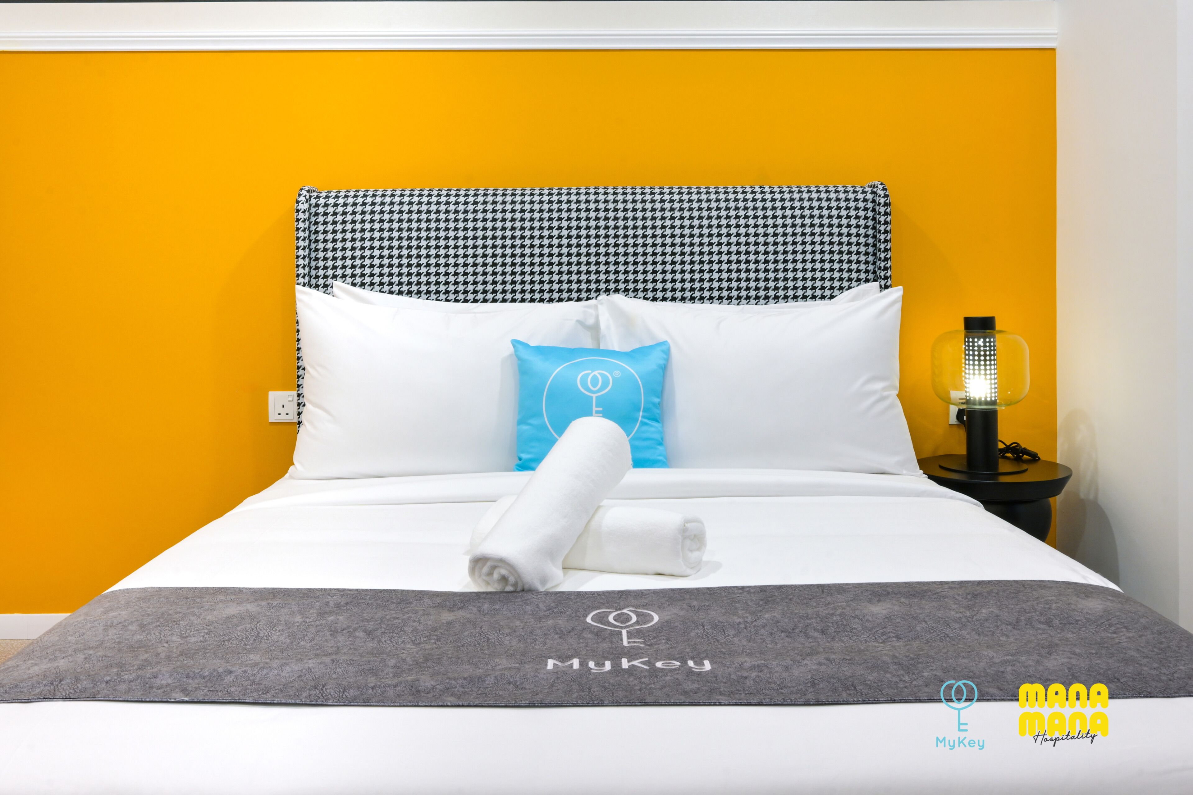 Una cama bien hecha con una colcha gris, almohadas blancas y una almohada decorativa azul. Una toalla blanca enrollada está colocada sobre la cama. El cabecero es a cuadros blancos y negros estilo pied de poule, y hay una mesita de noche con una lámpara al lado de la cama.