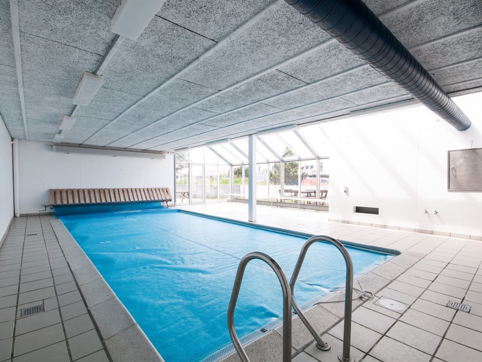Una piscina interior con paredes de vidrio transparente, un banco y suelo de baldosas.