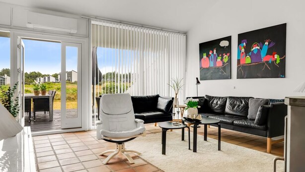 A modern living room with a black leather sofa, a white armchair, and a coffee table. There are two paintings on the wall and a sliding glass door leading to an outdoor area. - "Nis" - 900m from the sea by Interhome (Blokhus)
