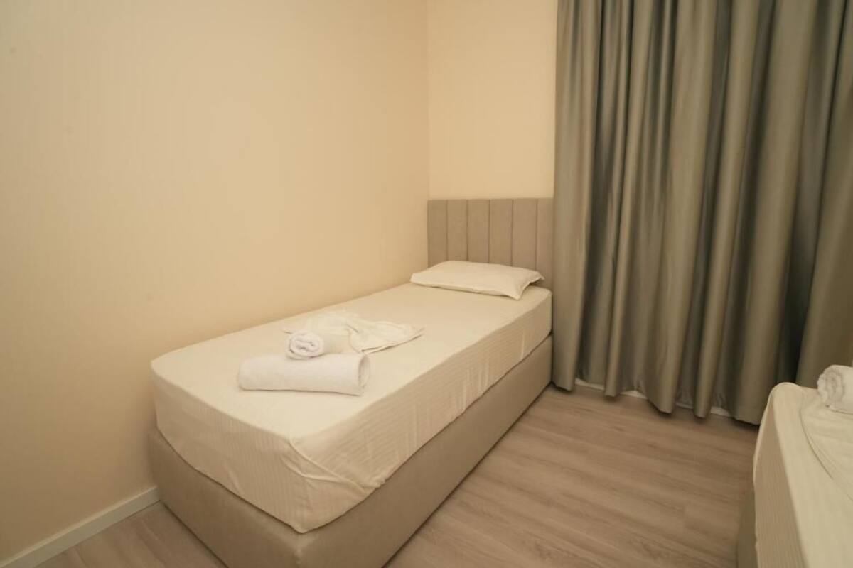 Un letto piccolo e ben rifatto con materasso e cuscino bianchi, un asciugamano piegato sul letto e una tenda parzialmente socchiusa.