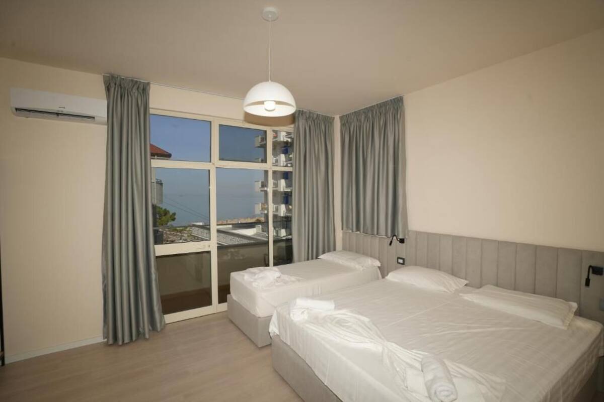 Una camera d'albergo con due letti, un'ampia finestra con tende, una lampada a soffitto e un condizionatore d'aria.